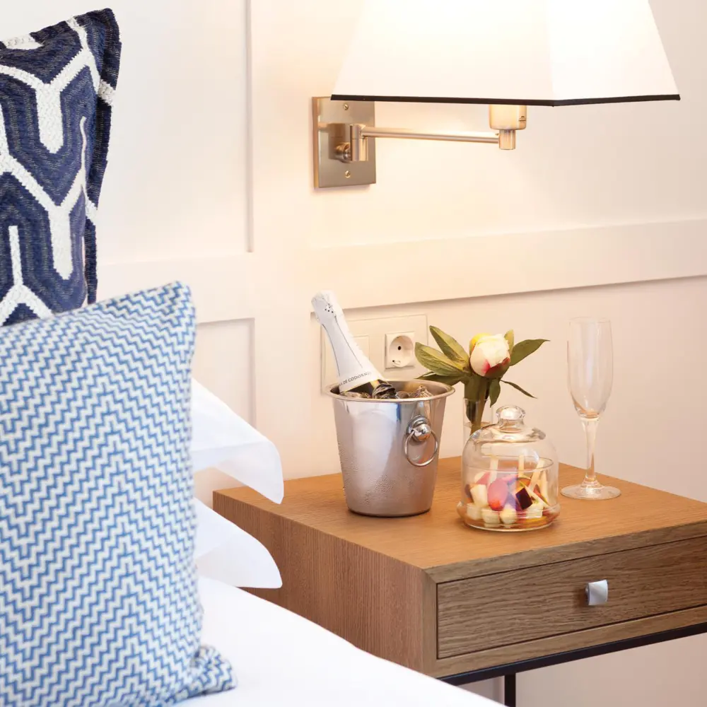 Cette table de chevet d'hôtel moderne en bois propose une bouteille de champagne au frais, une flûte en verre, un pot à confiseries ainsi que des oreillers aux motifs géométriques bleus et blancs.